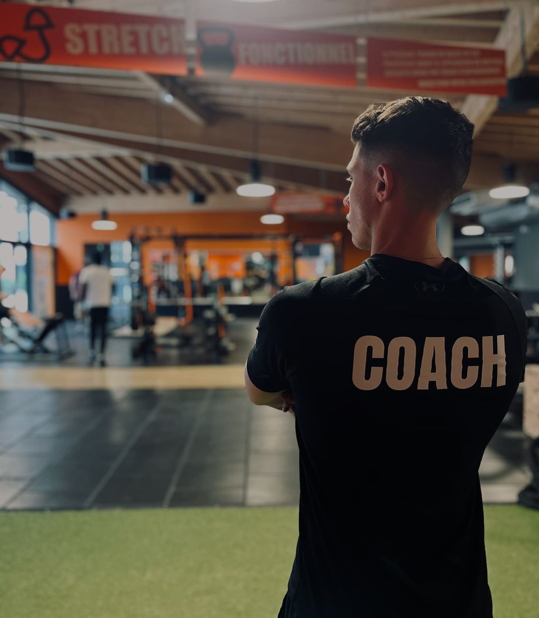 Coach de dos en t-shirt noir floqué COACH, salle de sport en arrière-plan flou